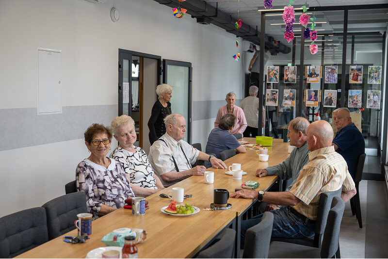 Gmina Baranów Ilustracja ukazuje wesołych seniorów – kobiety i mężczyźni, którzy siedzą przy stole i piją kawę, w nowoczesnej przestrzeni. Autorem zdjęcia jest Maciej Motylewski.