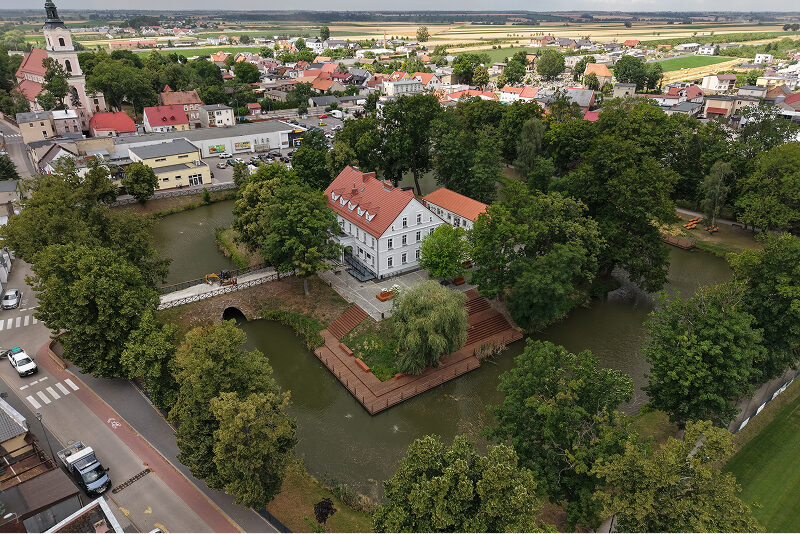 Gmina Krobia Zdjęcie z lotu ptaka ukazuje zabytkowy gmach z pochyłym dachem, znajdujący się na wyspie otoczonej fosą. Prowadzi na nią most. Wokół znajduje się miejska zabudowa i drzewa. Autorem fotografii jest Maciej Motylewski.