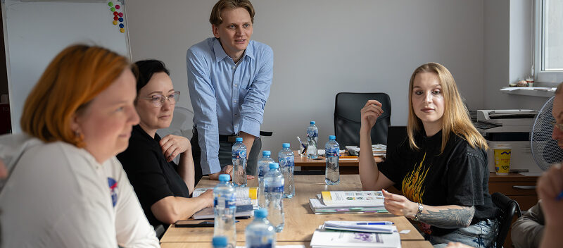 Fundacja Akme Fotografia przedstawia kilka osób, siedzących przy stole i jednego mężczyznę, który stoi i opiera się o stół. Na blacie leżą notatniki, stoją też butelki z wodą mineralną. Autorem zdjęcia jest Maciej Motylewski.