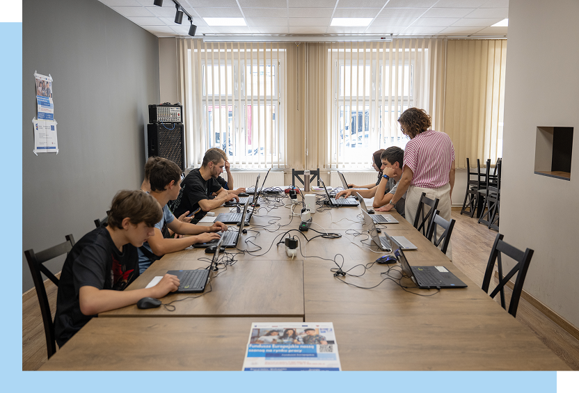 Fotografia przedstawia grupę młodzieży, która w skupieniu pracuje na laptopach rozłożonych na stolikach. Nadzoruje ich nauczycielka. Autorem zdjęcia jest Maciej Motylewski.