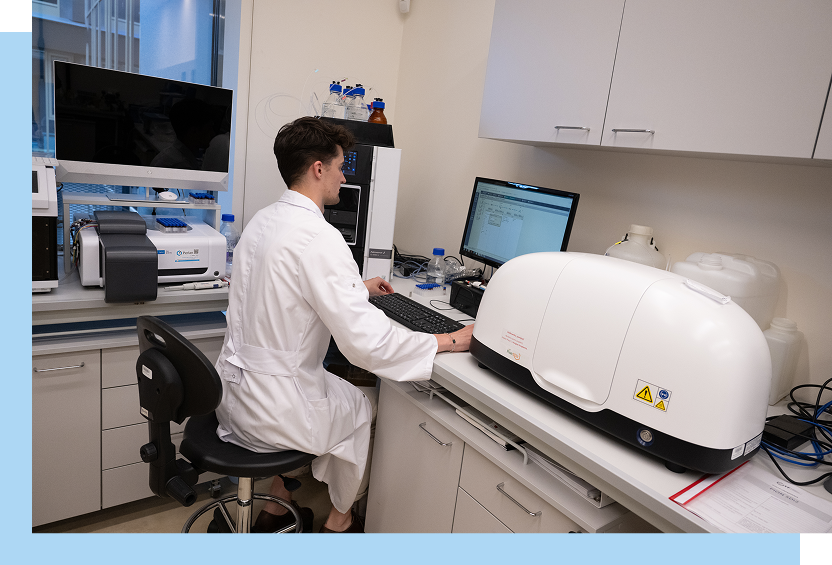 Zdjęcie przedstawia młodego mężczyznę w laboratoryjnym fartuchu, który siedzi przy komputerze. Wokół znajduje się sprzęt laboratoryjny. Autorem fotografii jest Maciej Motylewski.