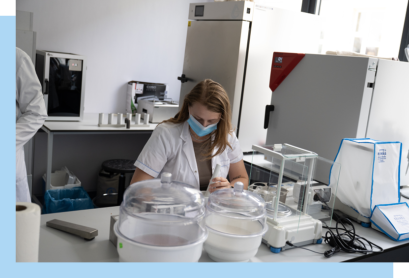 Scena ma miejsce w laboratorium. Widoczna jest kobieta, w ochronnej maseczce, która siedzi przy biurku i wykonuje badanie. Wokół niej znajduje się sprzęt laboratoryjny. Autorem zdjęcia jest Maciej Motylewski.