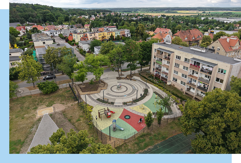 Fotografia z lotu ptaka przedstawia odnowiony plac w miejskiej przestrzeni, pomiędzy budynkami. Widać kolorową powierzchnię i sporo drzew. Autorem zdjęcia jest Maciej Motylewski.