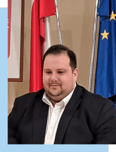 Na zdjęciu widoczny jest mężczyzna w garniturze. Za jego plecami znajdują się flagi Polski i Unii Europejskiej. Fotografia pochodzi z archiwum beneficjenta.