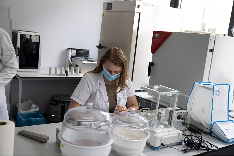 Scena ma miejsce w laboratorium. Widoczna jest kobieta, w ochronnej maseczce, która siedzi przy biurku i wykonuje badanie. Wokół niej znajduje się sprzęt laboratoryjny. Autorem zdjęcia jest Maciej Motylewski.