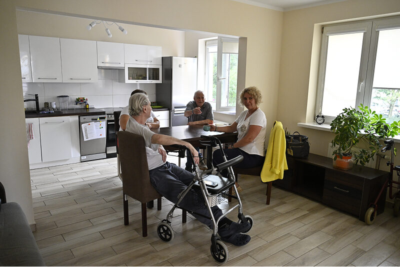 Zdjęcie przedstawia dwóch mężczyzn i dwie kobiety, którzy siedzą przy stole w kuchni, piją kawę i rozmawiają. Autorem fotografii jest Maciej Motylewski.