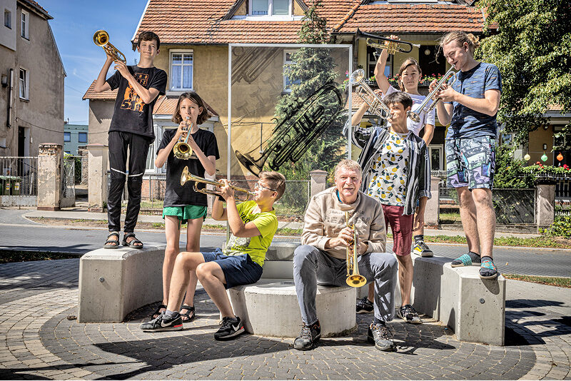 Ilustracja przedstawia grupę siedmioro muzyków pozujących do zdjęcia. Wśród nich są młodzi ludzie – dziewczynki i chłopcy oraz jeden starszy pan. Wszyscy trzymają trąbki. W tle widoczna jest także gablota z obrazkiem trąbki. Fotografia pochodzi z archiwum beneficjenta.