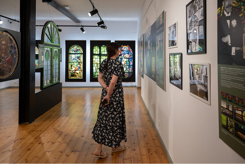 Na zdjęciu widoczna jest kobieta w długiej sukience, która przechadza się po wystawia muzealnej, przyglądając się eksponatom. Autorem fotografii jest Maciej Motylewski.