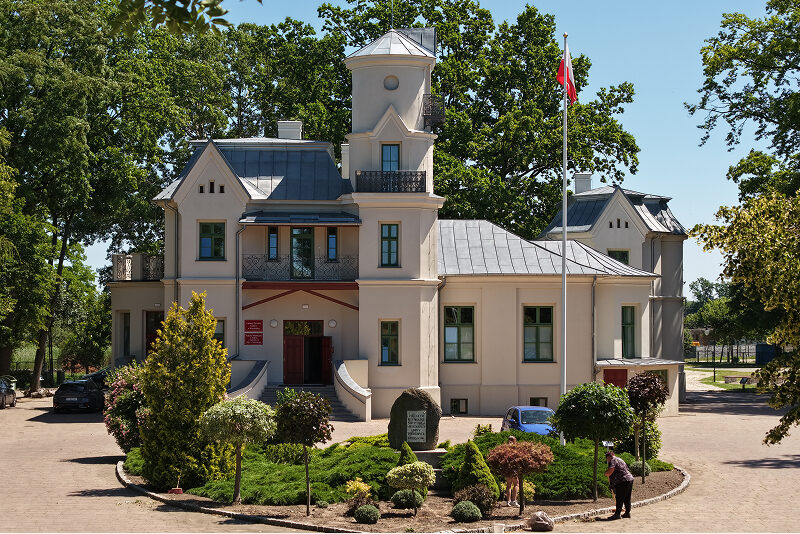 Zdjęcie przedstawia niewielki pałacyk, o nieregularnym kształcie. Przed nim znajduje się niewielki skwer z roślinami, w tle widoczne są zaś wysokie drzewa. Autorem fotografii jest Maciej Motylewski.