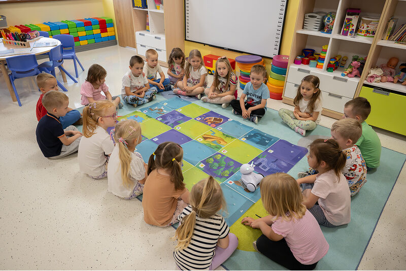 Fotografia przedstawia scenę w przedszkolu. Dzieci siedzą w kwadracie na dywanie i obserwują jeżdżącego mini robota. Autorem zdjęcia jest Maciej Motylewski.