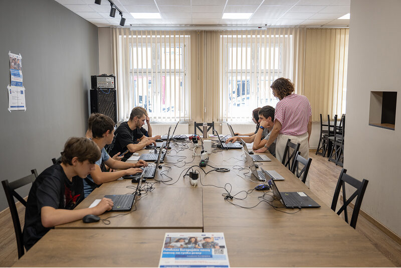 Fotografia przedstawia grupę młodzieży, która w skupieniu pracuje na laptopach rozłożonych na stolikach. Nadzoruje ich nauczycielka. Autorem zdjęcia jest Maciej Motylewski.