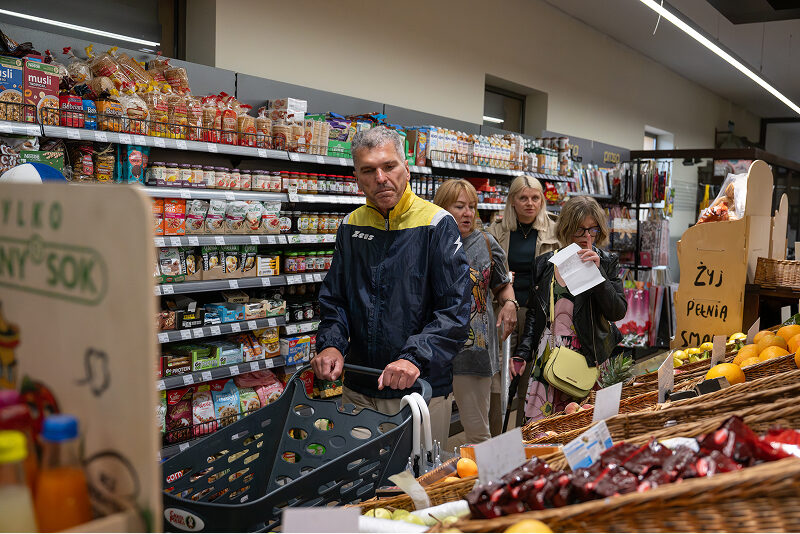 Zdjęcie przedstawia grupę czterech osób – mężczyzny, który pcha wózek sklepowy i trzech kobiet. Robią zakupy. Widoczne są regały pełne towaru. Autorem fotografii jest Maciej Motylewski.
