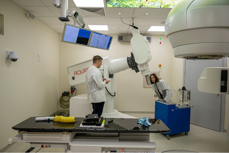 Zdjęcie wykonano w sali z wielkim medycznym robotem, służącym do napromieniania. Obsługują go kobieta i mężczyzna, oboje w lekarskich fartuchach. Autorem fotografii jest Maciej Motylewski.