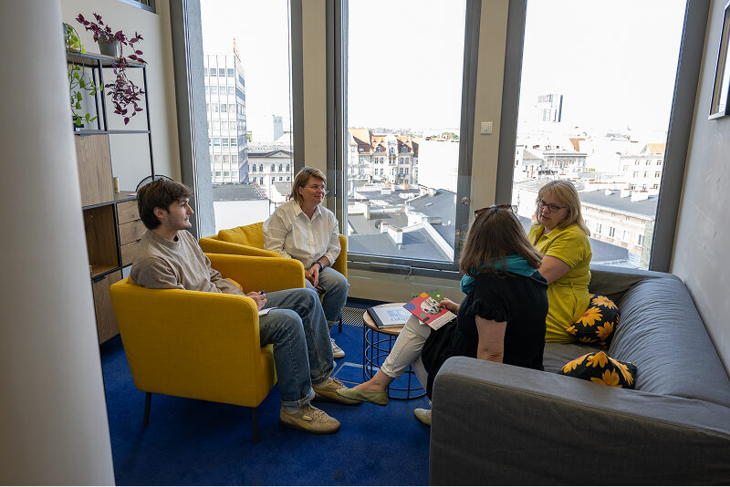 Zdjęcie przedstawia trzy dorosłe kobiety i jednego młodego mężczyznę, który siedzą przy małym okrągłym stoliku i rozmawiają. Za szybą widoczna jest panorama miasta. Autorem fotografii jest Maciej Motylewski.
