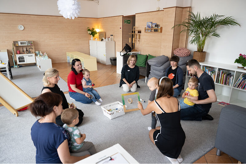 Zdjęcie przedstawia opiekunów – młodych rodziców, wśród których są mamy i jeden tata, ze swoimi dziećmi. Siedzą na dywanie i grają w grę. Autorem fotografii jest Maciej Motylewski.