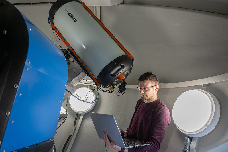 Zdjęcie wykonano we wnętrzu ogromnej kopuły, która jest obserwatorium astronomicznym. Widoczny jest mężczyzna w okularach, trzymający laptopa. Stoi obok urządzenia przypominającego teleskop. Autorem fotografii jest Maciej Motylewski.