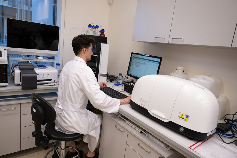 Zdjęcie przedstawia młodego mężczyznę w laboratoryjnym fartuchu, który siedzi przy komputerze. Wokół znajduje się sprzęt laboratoryjny. Autorem fotografii jest Maciej Motylewski.