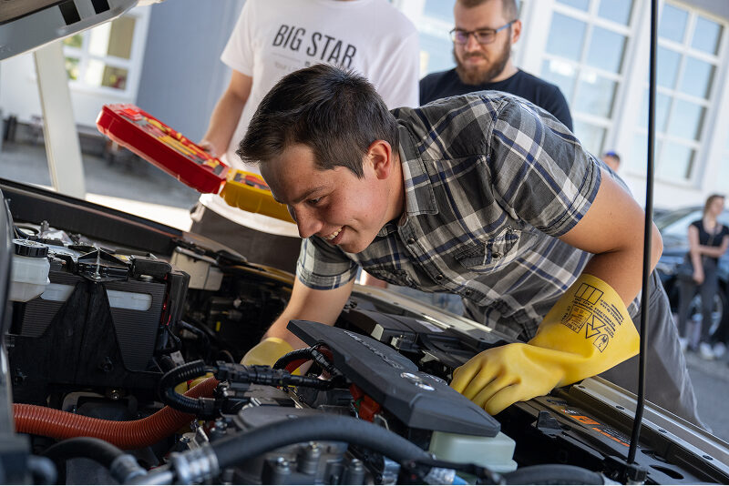 Na zdjęciu widać młodego, uśmiechniętego mężczyznę, który zagląda pod maskę elektrycznego samochodu. Na rękach ma gumowe rękawice. Autorem fotografii jest Maciej Motylewski.