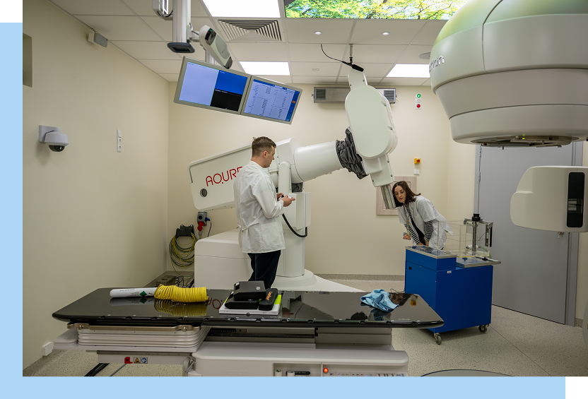 Zdjęcie wykonano w sali z wielkim medycznym robotem, służącym do napromieniania. Obsługują go kobieta i mężczyzna, oboje w lekarskich fartuchach. Autorem fotografii jest Maciej Motylewski.