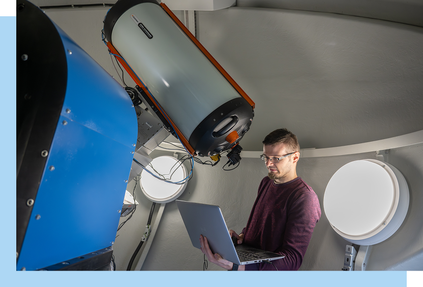 Politechnika Poznańska Zdjęcie wykonano we wnętrzu ogromnej kopuły, która jest obserwatorium astronomicznym. Widoczny jest mężczyzna w okularach, trzymający laptopa. Stoi obok urządzenia przypominającego teleskop. Autorem fotografii jest Maciej Motylewski.