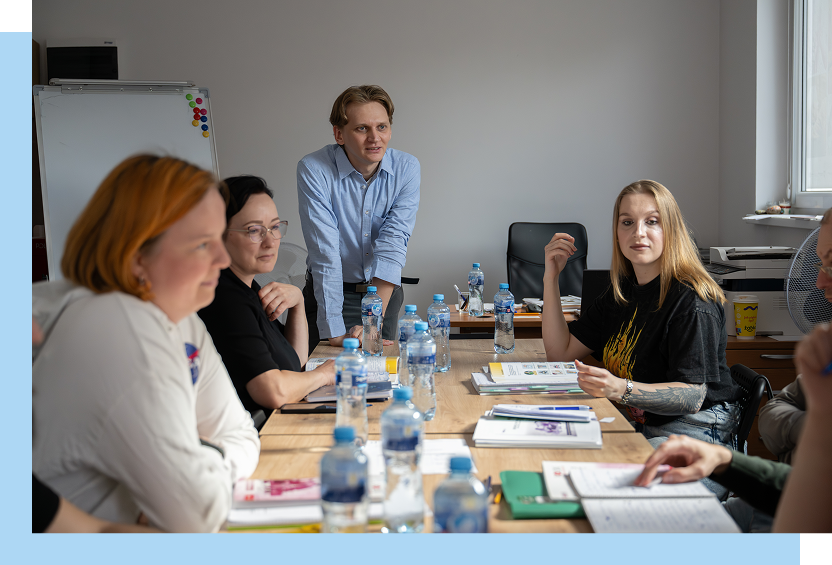 Fundacja Akme Fotografia przedstawia kilka osób, siedzących przy stole i jednego mężczyznę, który stoi i opiera się o stół. Na blacie leżą notatniki, stoją też butelki z wodą mineralną. Autorem zdjęcia jest Maciej Motylewski.