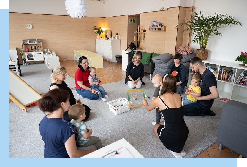 Gmina Czarnków/Centrum Usług Społecznych Zdjęcie przedstawia opiekunów – młodych rodziców, wśród których są mamy i jeden tata, ze swoimi dziećmi. Siedzą na dywanie i grają w grę. Autorem fotografii jest Maciej Motylewski.