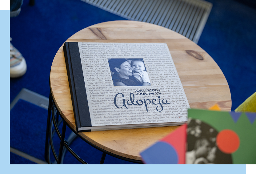Regionalny Ośrodek Polityki Społecznej w Poznaniu Na fotografii, na okrągłym drewnianym stoliku leży książka, zatytułowana: Album rodzin adopcyjnych. Adopcja. Na okładce, utrzymanej w beżowym odcieniu, widoczne jest też zdjęcie uśmiechniętej mamy z niemowlakiem. Autorem zdjęcia jest Maciej Motylewski.