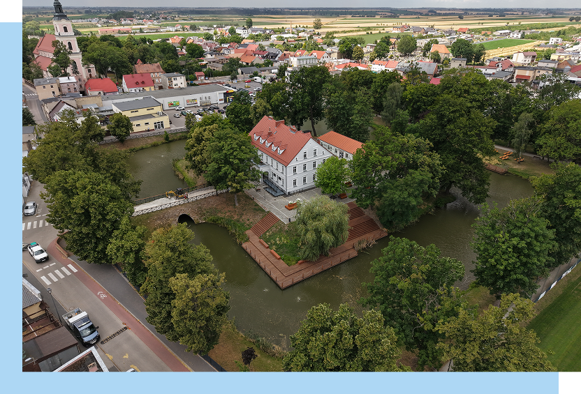 Gmina Krobia Zdjęcie z lotu ptaka ukazuje zabytkowy gmach z pochyłym dachem, znajdujący się na wyspie otoczonej fosą. Prowadzi na nią most. Wokół znajduje się miejska zabudowa i drzewa. Autorem fotografii jest Maciej Motylewski.