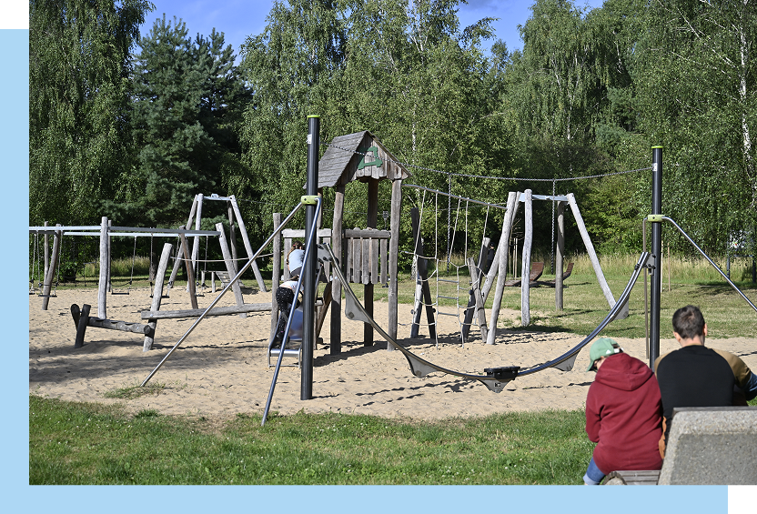 Zdjęcie ukazuje plac zabaw na piaszczystym podłożu. Widoczne są urządzenia zabawowe z drewna.