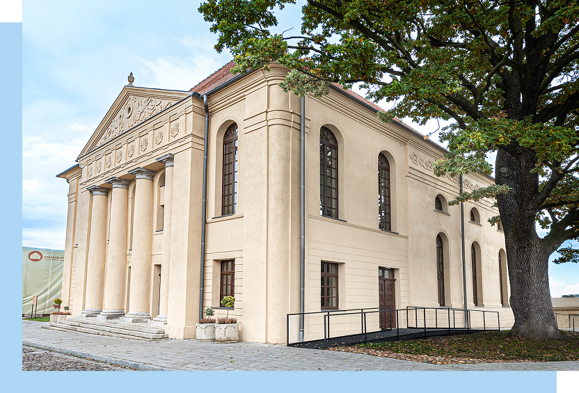 Zdjęcie ukazuje odnowiony budynek synagogi, do której prowadzi brukowana droga. Obok budynku rośnie rozłożyste drzewo.