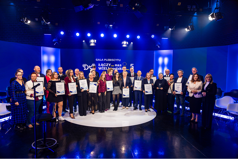 1 Na zdjęciu znajduje się grupa kilkudziesięciu osób, elegancko ubranych, które stoją na scenie i pozują do wspólnej fotografii. Scena ma miejsce w studio telewizyjnym, widoczne są więc niebieskie ekrany, kamery i oświetlenie. Zdjęcie pochodzi z archiwum Urzędu Marszałkowskiego Województwa Wielkopolskiego.